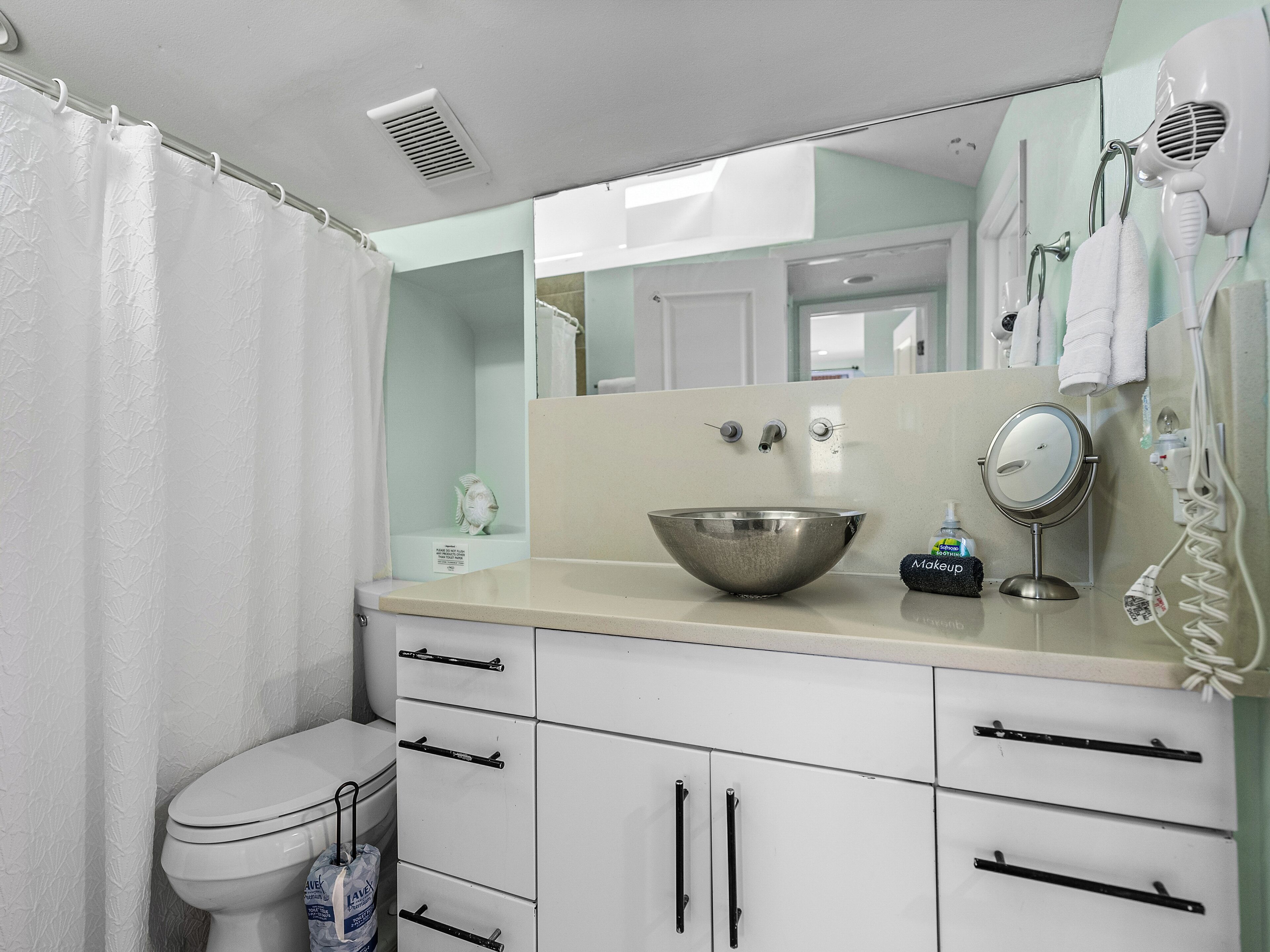 A bathroom with a white toilet, a stainless steel sink, a mirror, and a hairdryer on the wall.