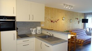 A compact kitchen with a built-in oven, a coffee maker, and a sink. There is a wooden wall paneling and a small seating area with a plaid sofa.