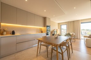 Cozinha moderna com eletrodomésticos integrados, área de jantar com mesa e cadeiras de madeira, e sala de estar com sofá e grandes janelas.