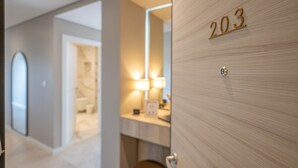 A hotel room with a wooden door marked '203', a bathroom with a mirror, and a hallway leading to another room.