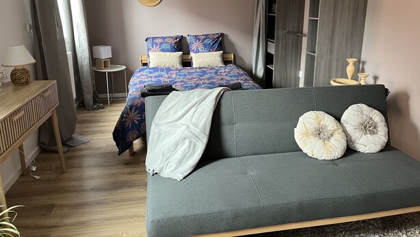 A modern bedroom with a grey sofa, a bed with floral bedding, a wooden nightstand, and a wardrobe. - Charmant Studio Proche Centre Ambiance Cosy (Beauvais)