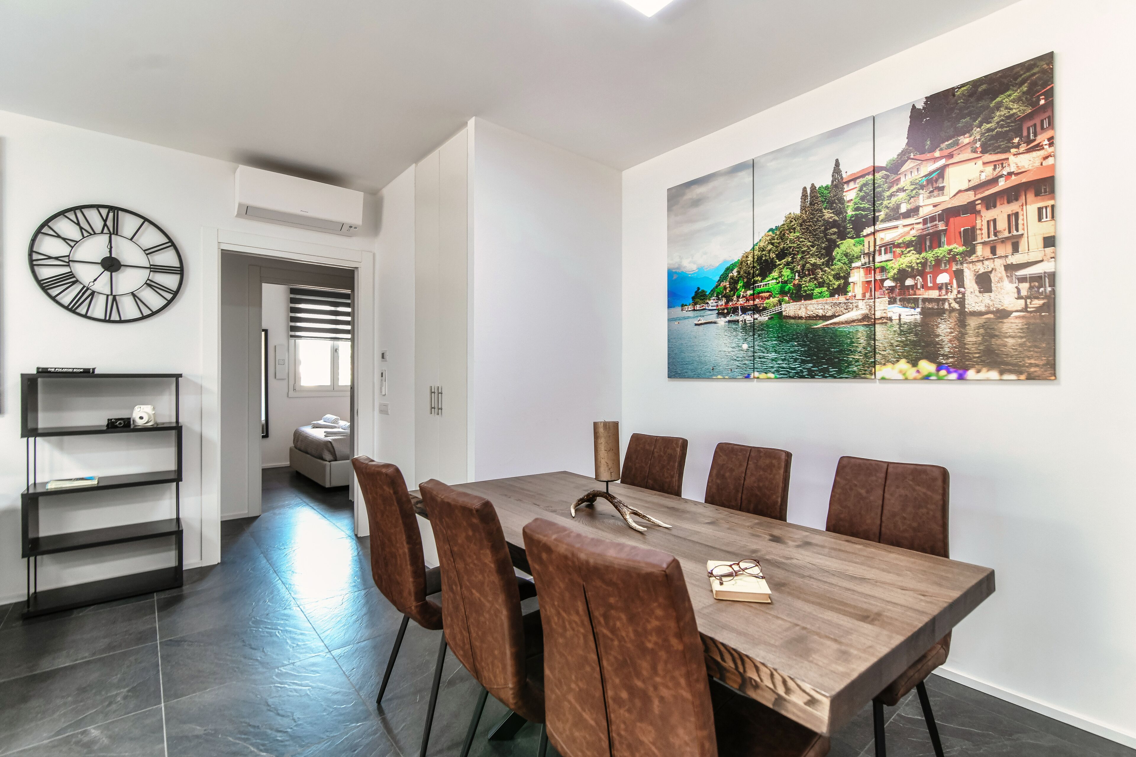 A dining area with a wooden table, brown upholstered chairs, and a large painting of a coastal town.