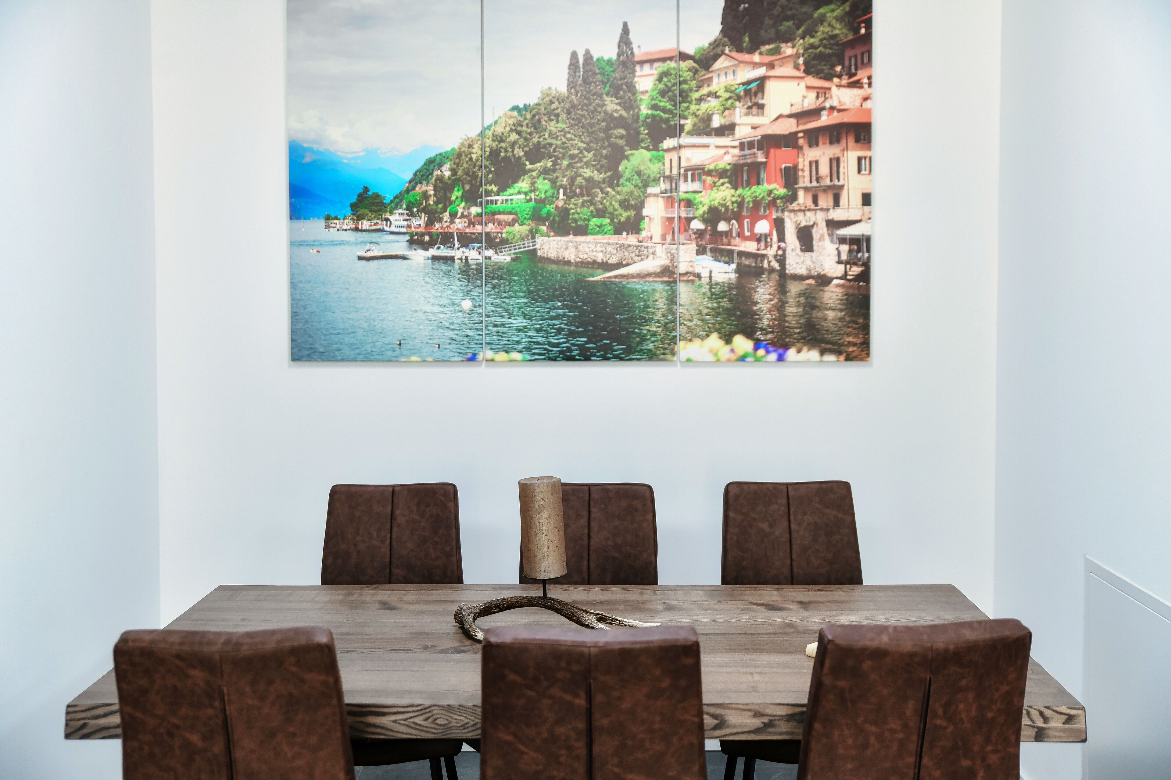 Un coin repas avec une table en bois et six chaises marron, et sur le mur derrière, un grand tableau représentant un paysage pittoresque d’une ville côtière.