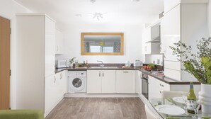 A modern kitchen with white cabinets, a built-in washing machine, a dining area with a glass table, and a ceiling fan. - 17 Astor Court, Newquay -  an apartment that sleeps 4 guests  in 2 bedrooms (Newquay)