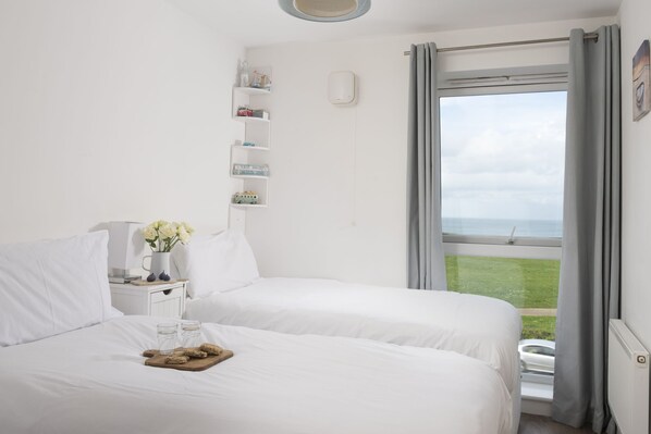 A bedroom with two beds, a bedside table with a vase of flowers, and a view of the sea through a large window.
