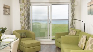 A living room with a glass door leading to a balcony with a view of the sea. There is a green sofa, a small green armchair, and a glass dining table with a white plate and cutlery. - 17 Astor Court, Newquay -  an apartment that sleeps 4 guests  in 2 bedrooms (Newquay)