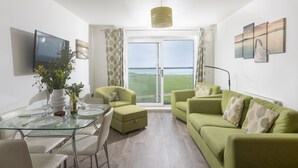 A modern living room with a glass dining table, green sofas, and a large window with a view of the sea. - 17 Astor Court, Newquay -  an apartment that sleeps 4 guests  in 2 bedrooms (Newquay)