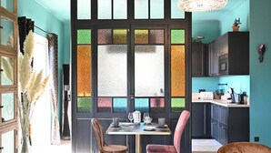 A modern kitchen with dark cabinets, a dining table with chairs, and a large window with colorful glass panes. - Alexandre Chambre D'hôte (LA LONDE LES MAURES)