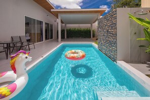 A swimming pool with a unicorn float and a ring float, surrounded by a modern patio area with a covered seating space and a stone wall.