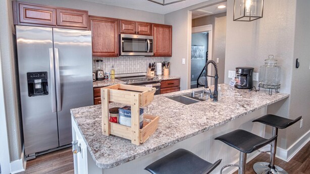 A modern kitchen with stainless steel appliances, wooden cabinets, granite countertops, and bar stools. - Lovely 3 BR Home Only 9 Minutes From Downtown (San Antonio)