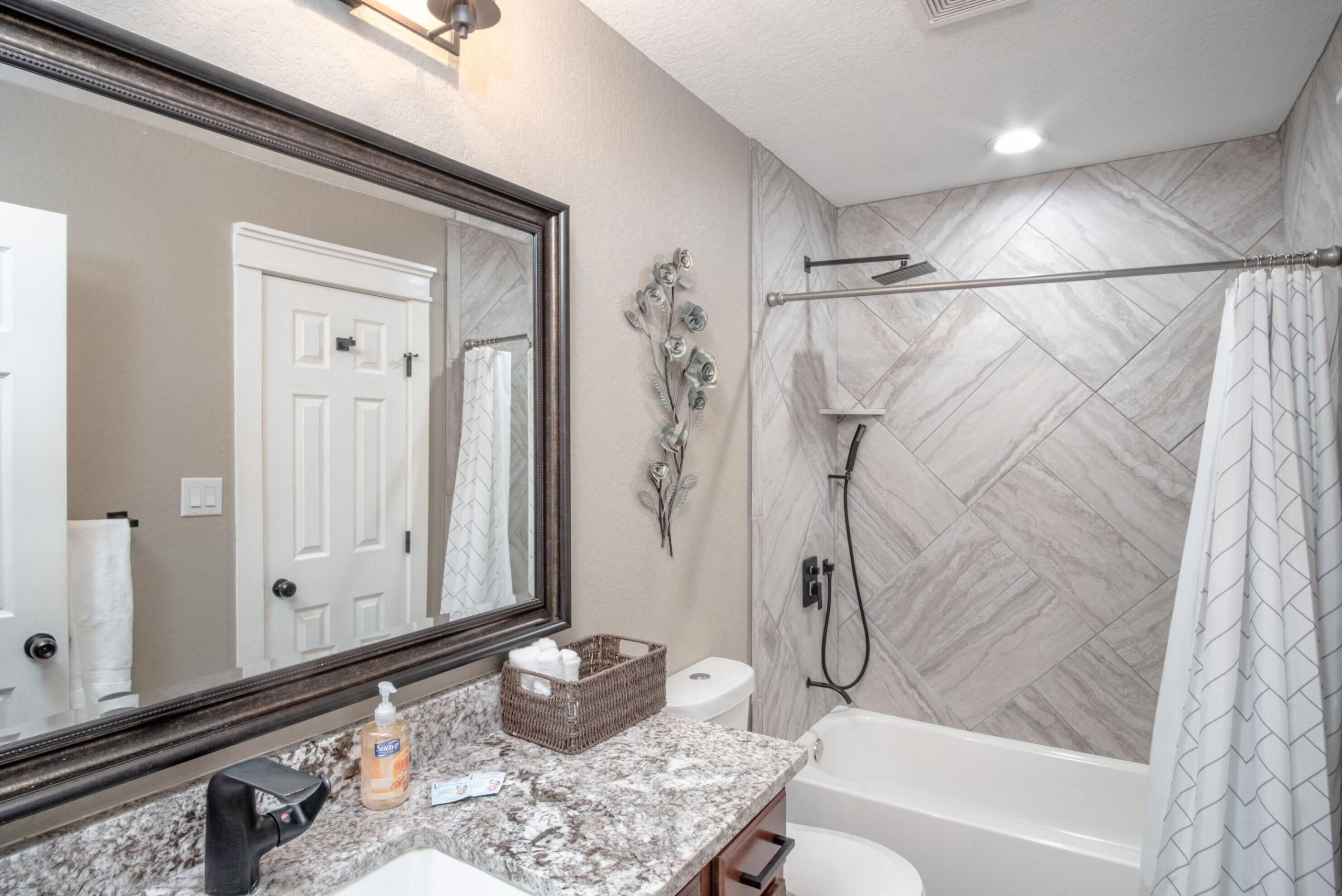 A bathroom with a marble countertop, a large mirror, a shower with a herringbone tile pattern, and a white door.