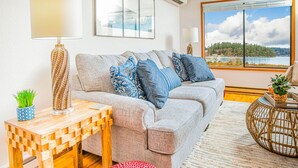 A living room with a grey sofa, blue and white patterned cushions, a wooden coffee table, a red ottoman, and a large window with a view of a lake and trees. - Cornet Bay Cottage in Oak Harbor (Oak Harbor)