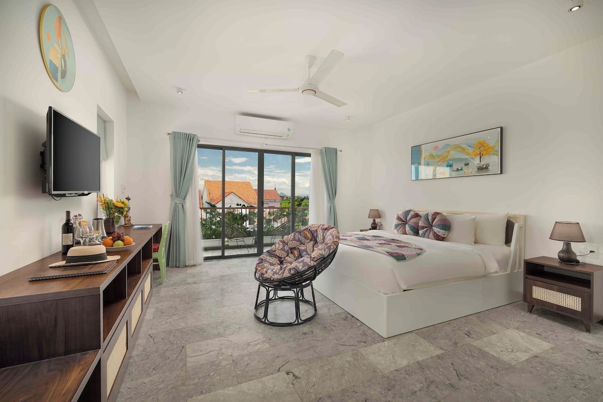 A modern bedroom with a large bed, a flat-screen TV, a ceiling fan, and a balcony with a view of houses.