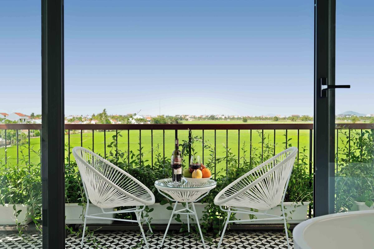 A balcony with two white chairs, a small table with a bottle and glasses, and a bowl of fruit.
