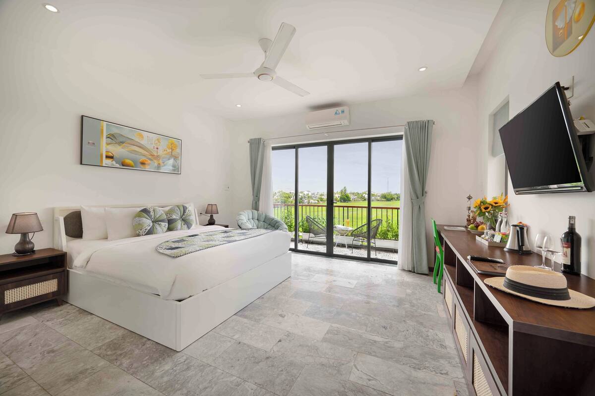 A bedroom with a bed, bedside tables, a TV, a balcony with a view, and an air conditioning unit.