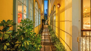 Un estrecho pasillo con piso de piedra, flanqueado por paredes amarillas y ventanas amplias, que conduce a una habitación con cama y vistas al cielo.