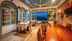 Un comedor con mesa y sillas de madera, ventanas amplias con marcos azules y vistas a la ciudad al anochecer.