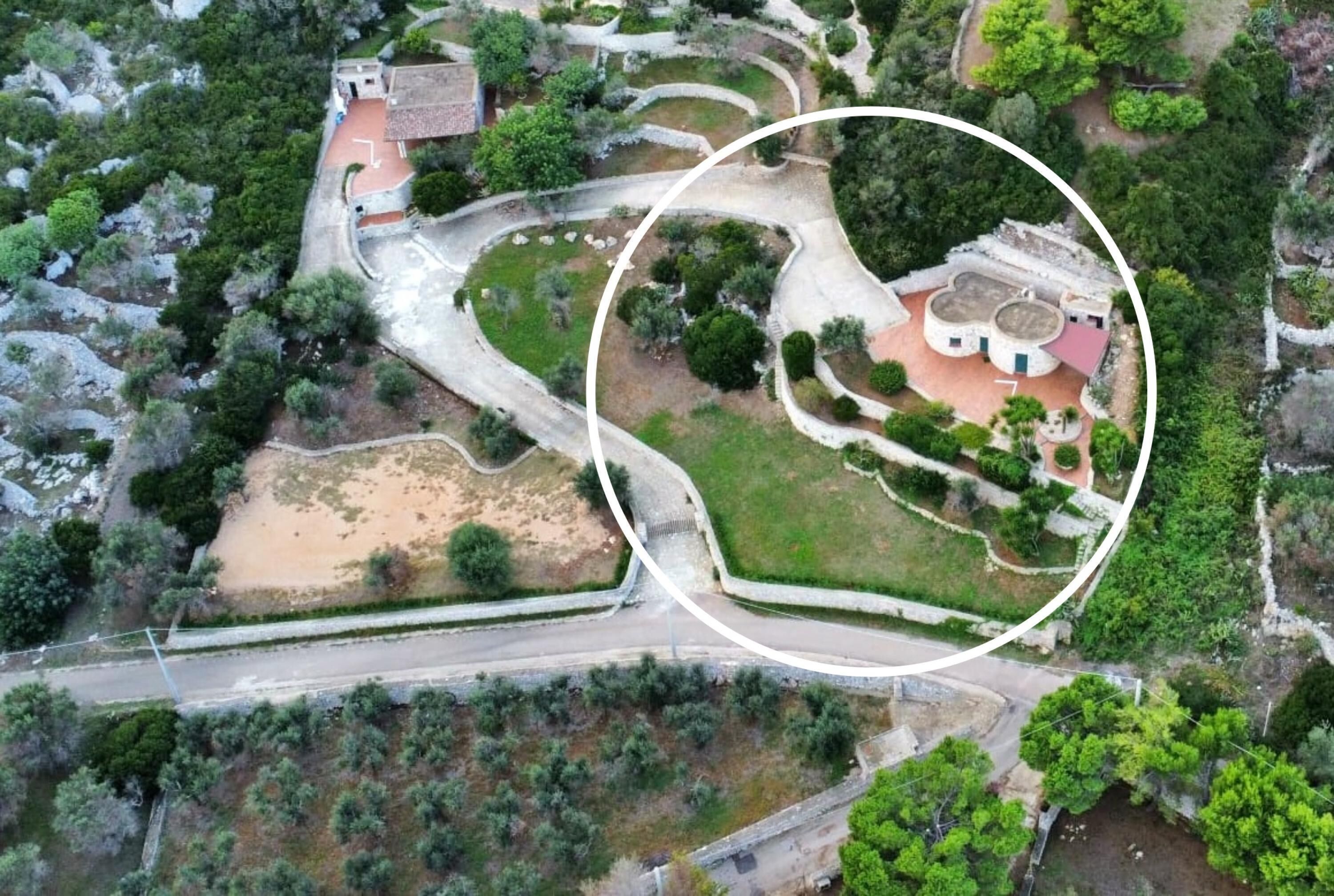 An aerial view of a landscaped area with a winding road, a small building with a red roof, and a circular garden area.