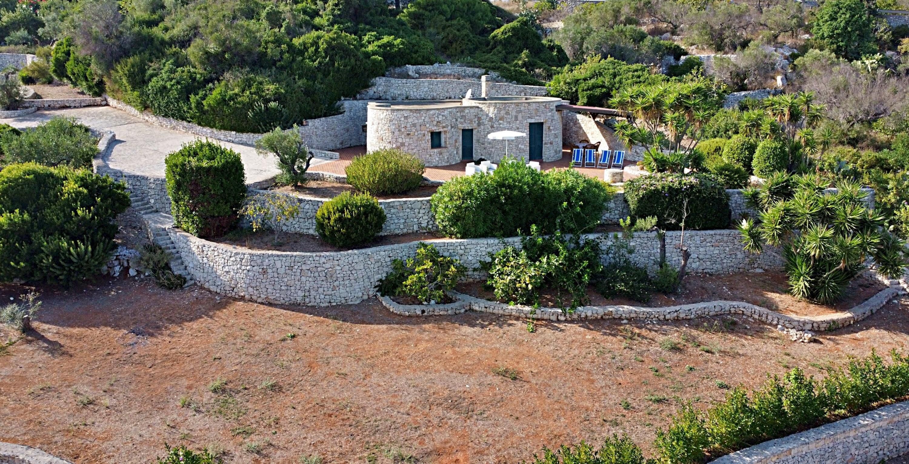 Une maison en pierre à plan circulaire, entourée d’un mur de pierre et d’une végétation luxuriante.