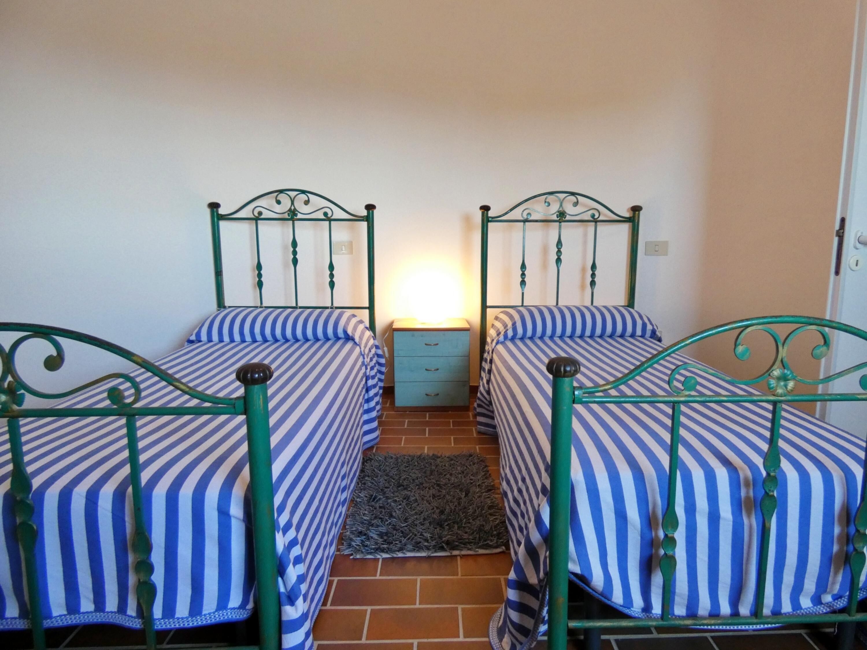 A room with two single beds, each with a striped bedspread, a small bedside table, and a rug on the floor.