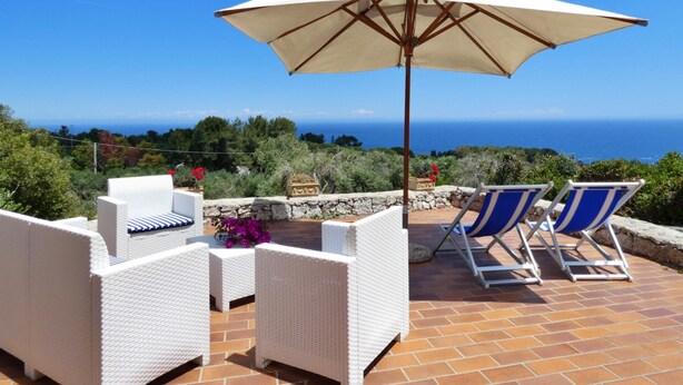 A patio with white wicker furniture, a large umbrella, and a view of the sea. - Villa Agapanto - Trullo Agap con Vista Mare e Giardino Privato (Tricase)