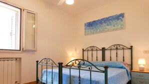 A bedroom with a bed, a nightstand, a radiator, a window, and a painting on the wall. - Villa Agapanto - Trullo Agap con Vista Mare e Giardino Privato (Tricase)