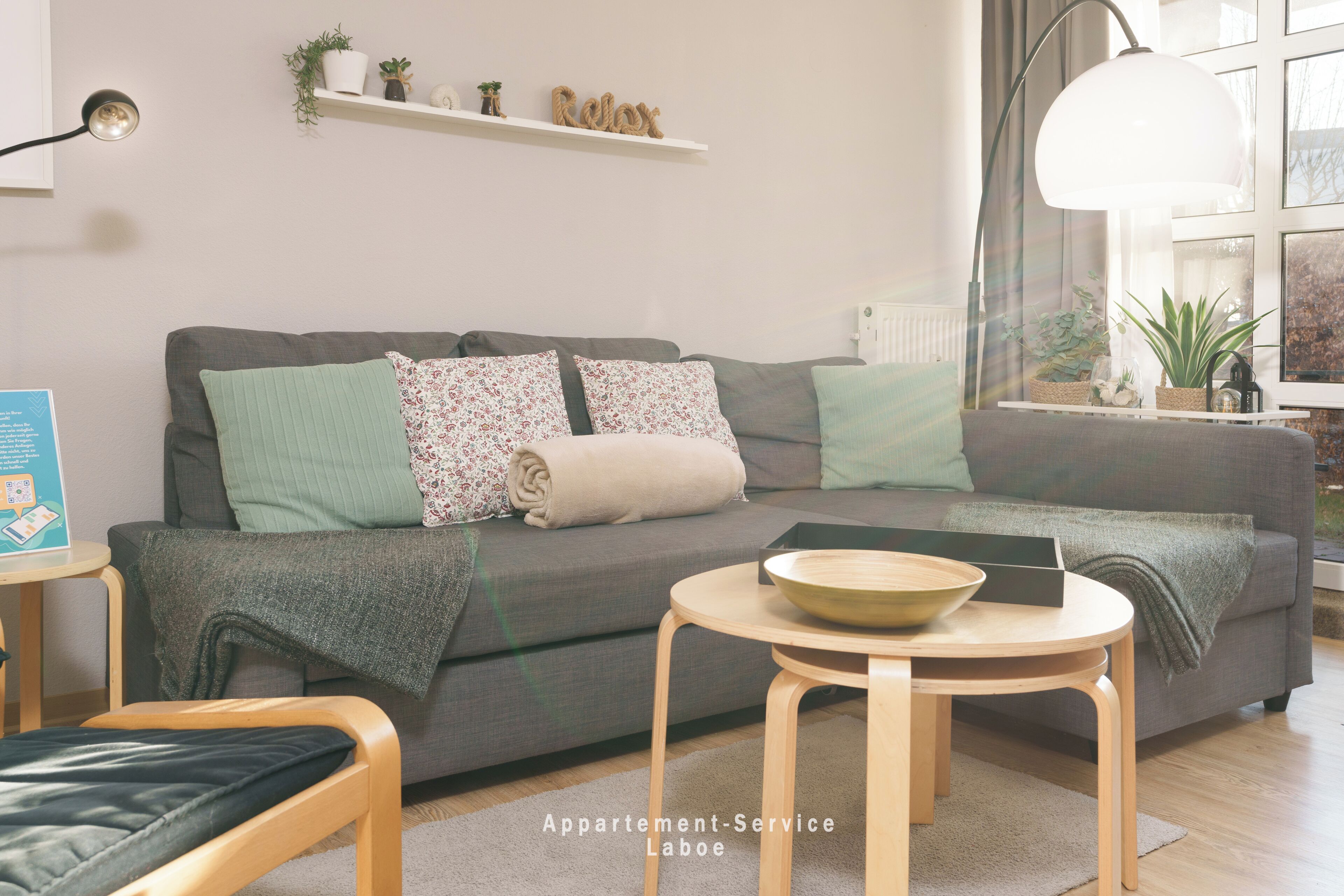 Ein modernes Wohnzimmer mit einer grauen Couch, einem Holztisch und einer Lampe. Regale mit Pflanzen und ein Schild mit der Aufschrift 'Relax'.