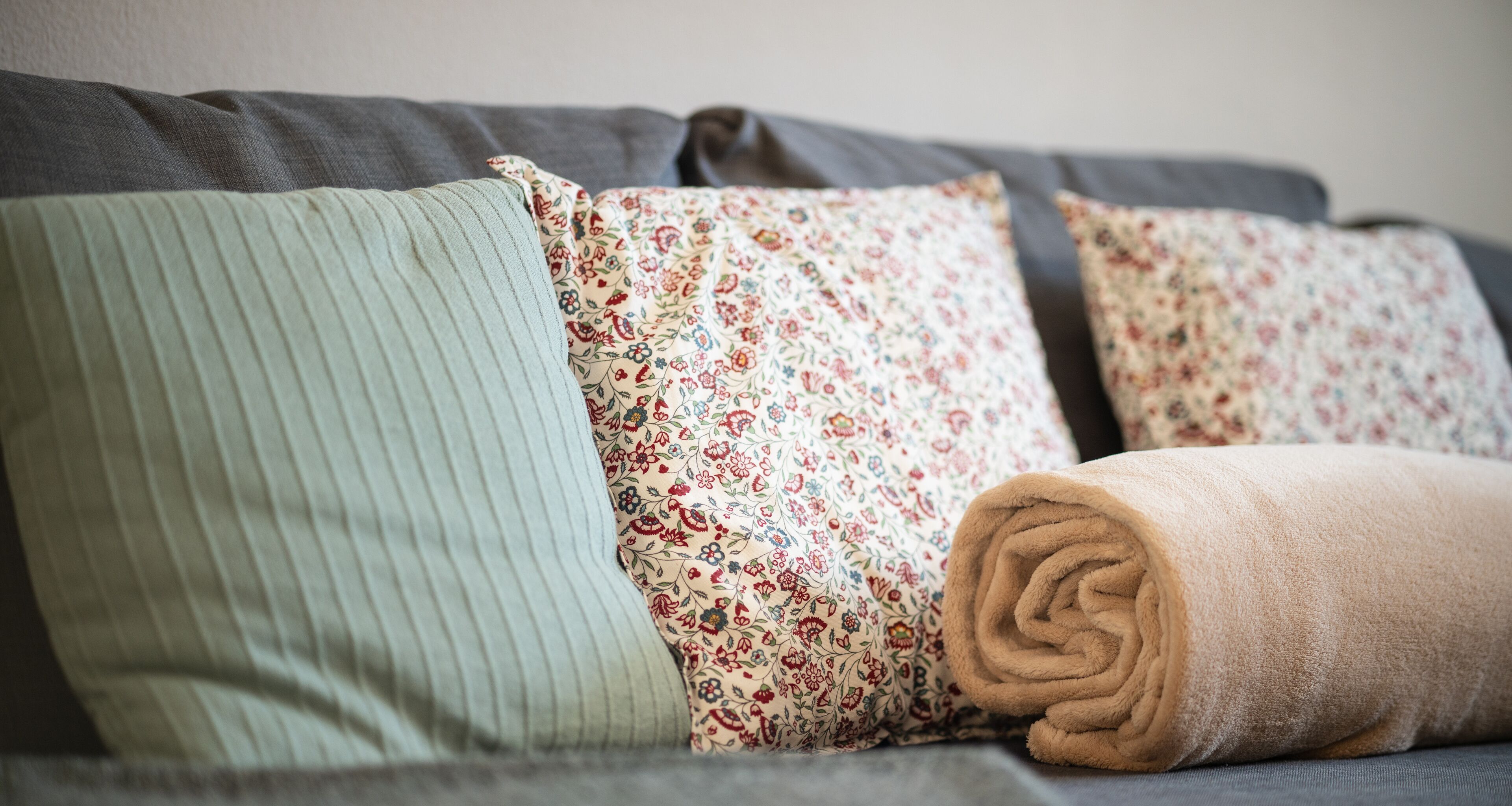 A grey sofa with three pillows: one striped, one floral, and one rolled-up beige towel.