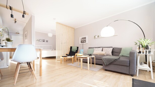 Un salon moderne avec un canapé gris, une table à manger en bois et une chaise blanche. On y trouve également une chaise noire avec un plaid vert, une table basse ronde et un lampadaire.