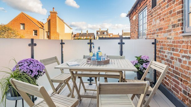 Une terrasse en bois avec une table et des chaises, entourée de murs en briques et offrant une vue sur des maisons.
