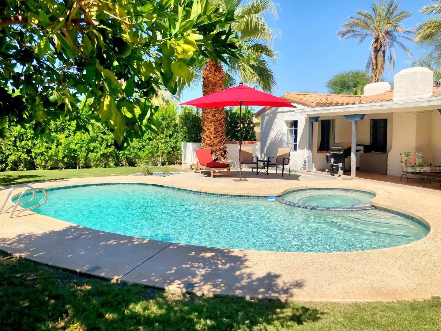 Ein Pool mit geschwungener Form, umgeben von einer Terrasse mit Gartenmöbeln und einem roten Sonnenschirm.