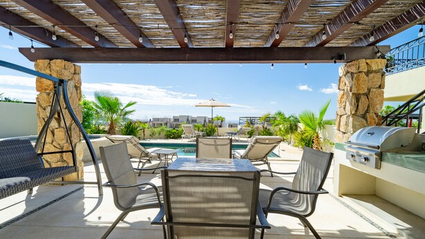 En udendørs patio med et bord, stole, grill og en pool i baggrunden.