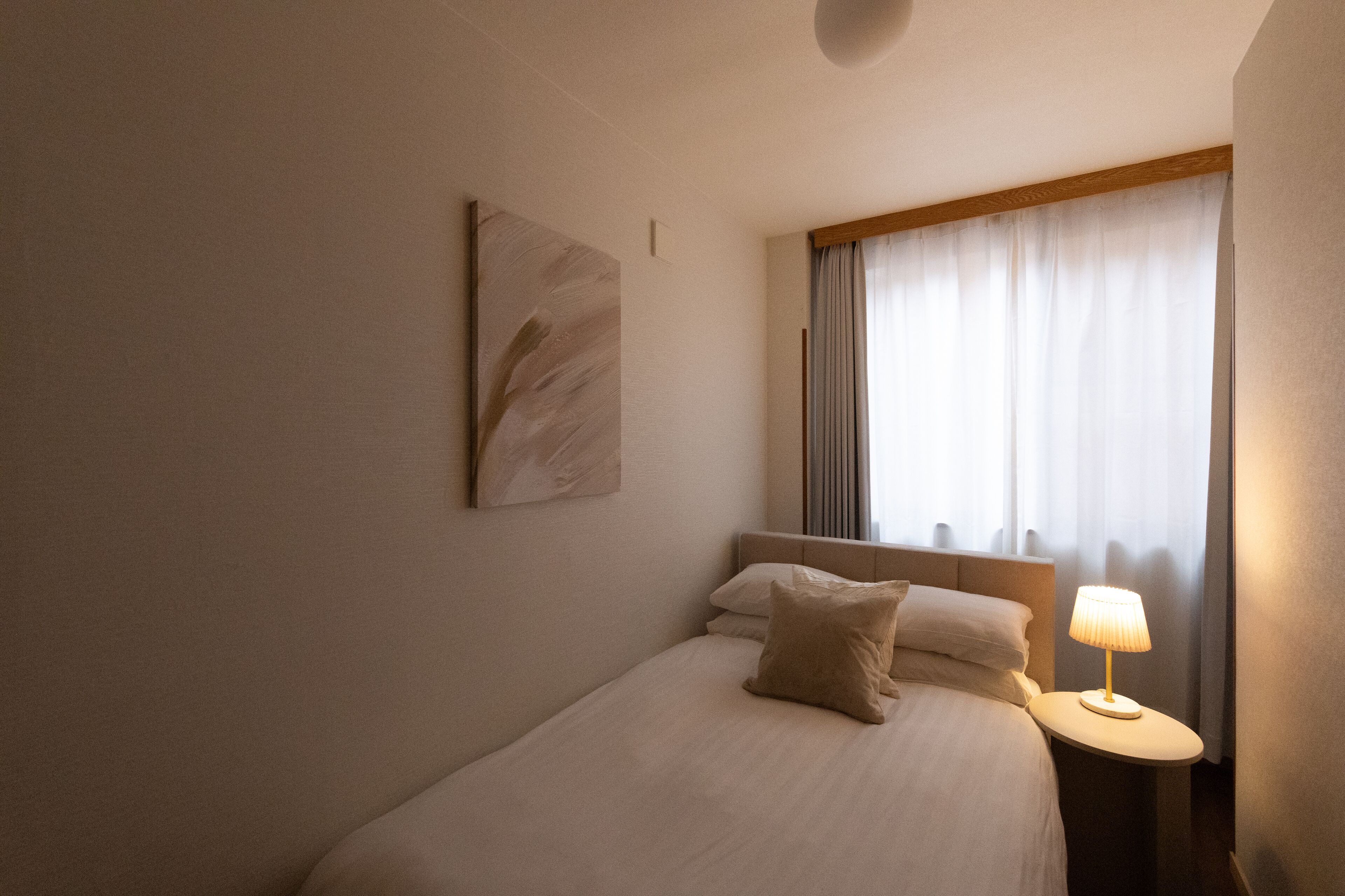 Ein Hotelzimmer mit einem Bett, einem Nachttisch mit Lampe, einem Fenster mit Vorhängen und einem Wandbild.