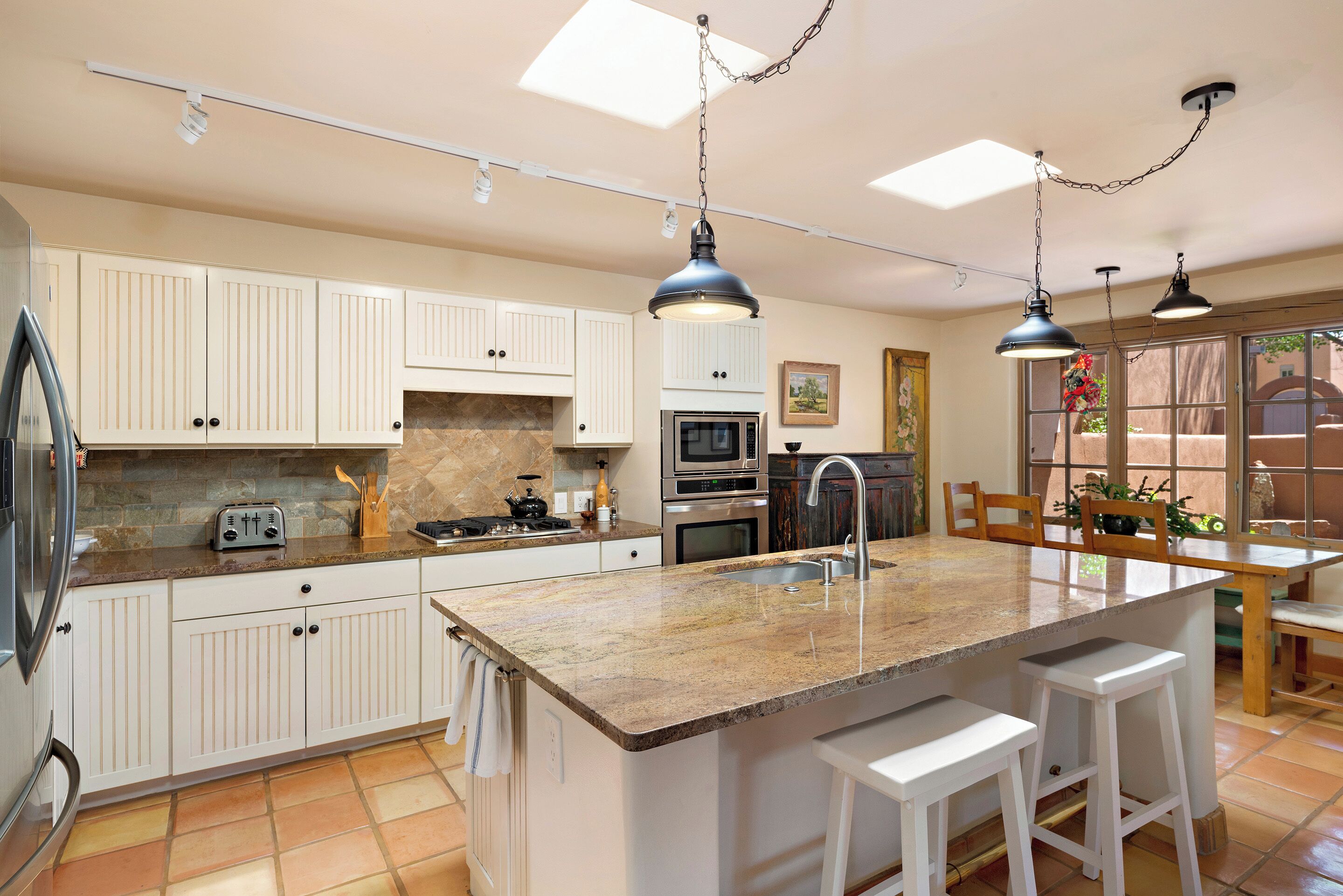 Una cocina con armarios blancos, encimera de granito, una isla central con taburetes de bar, un lavaplatos e iluminación colgante.
