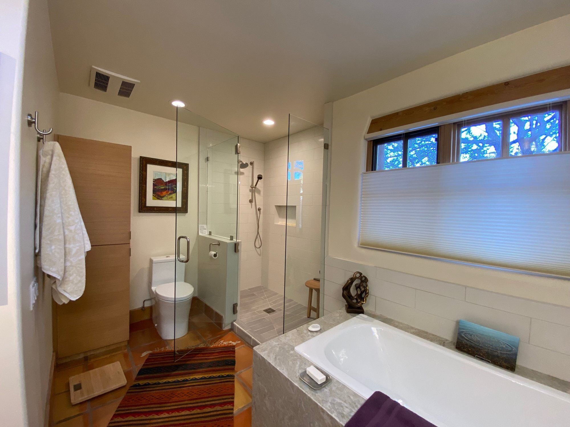A modern bathroom with a glass-enclosed shower, a white toilet, a bathtub, and a window with blinds.