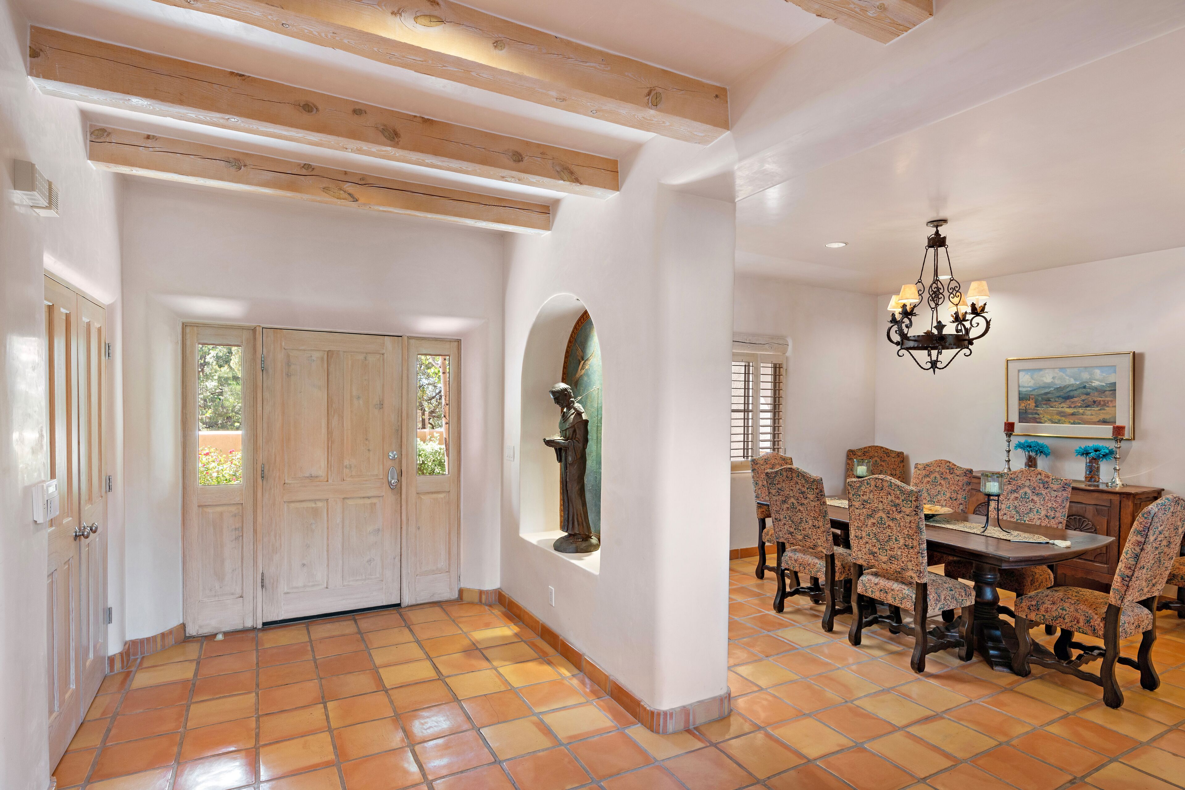 A spacious dining area with a large wooden table and patterned chairs, a chandelier, and a statue in a niche.