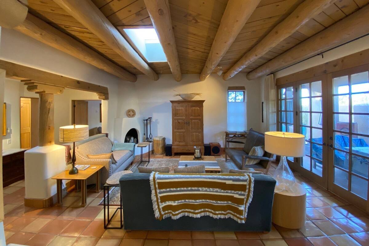 A spacious living room with a high ceiling, wooden beams, a fireplace, and large windows.