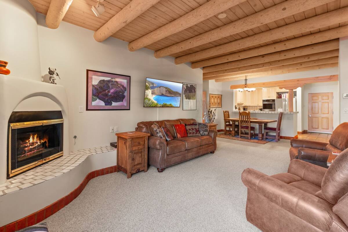 A living room with a fireplace, a sofa, a leather armchair, a dining area with a table and chairs, and a kitchen in the background.