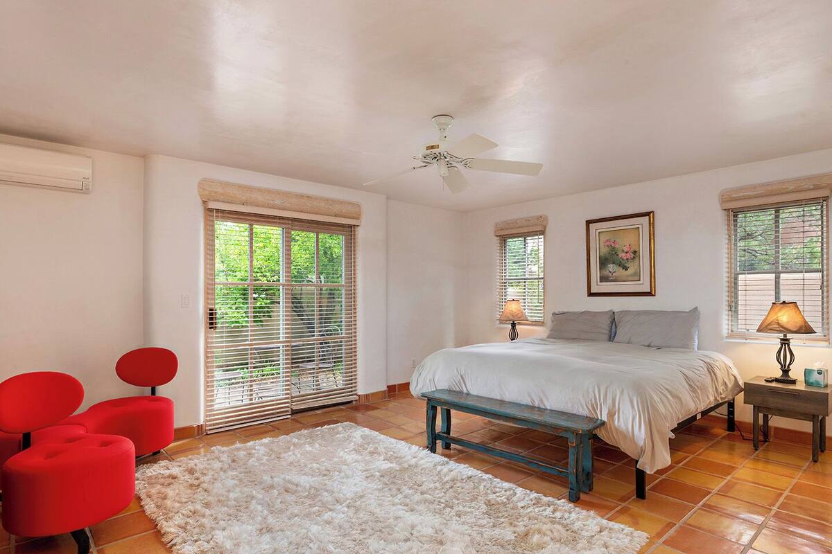 A bedroom with a bed, two bedside lamps, a ceiling fan, a window with blinds, a red chair, a bench, and a rug.