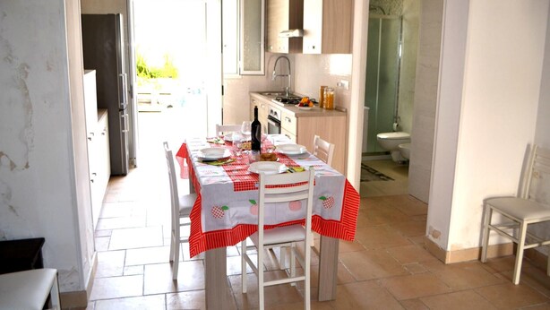 Un coin repas avec une table dressée pour quatre personnes, une cuisine à l’arrière-plan et une salle de bain visible à travers une porte ouverte.