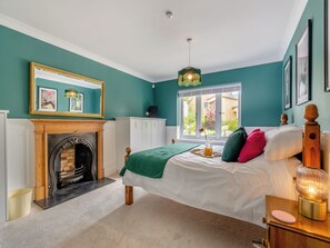 A bedroom with a fireplace, a bed with pillows, a bedside table with a lamp, and a window with a view of the outside.
