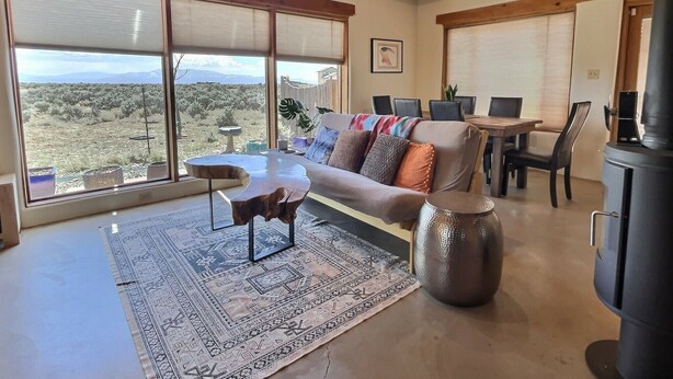 A spacious living room with a large window, a cozy sofa, a dining area, and a fireplace. - Adobe Tranquillo Mountain Views (El Prado)