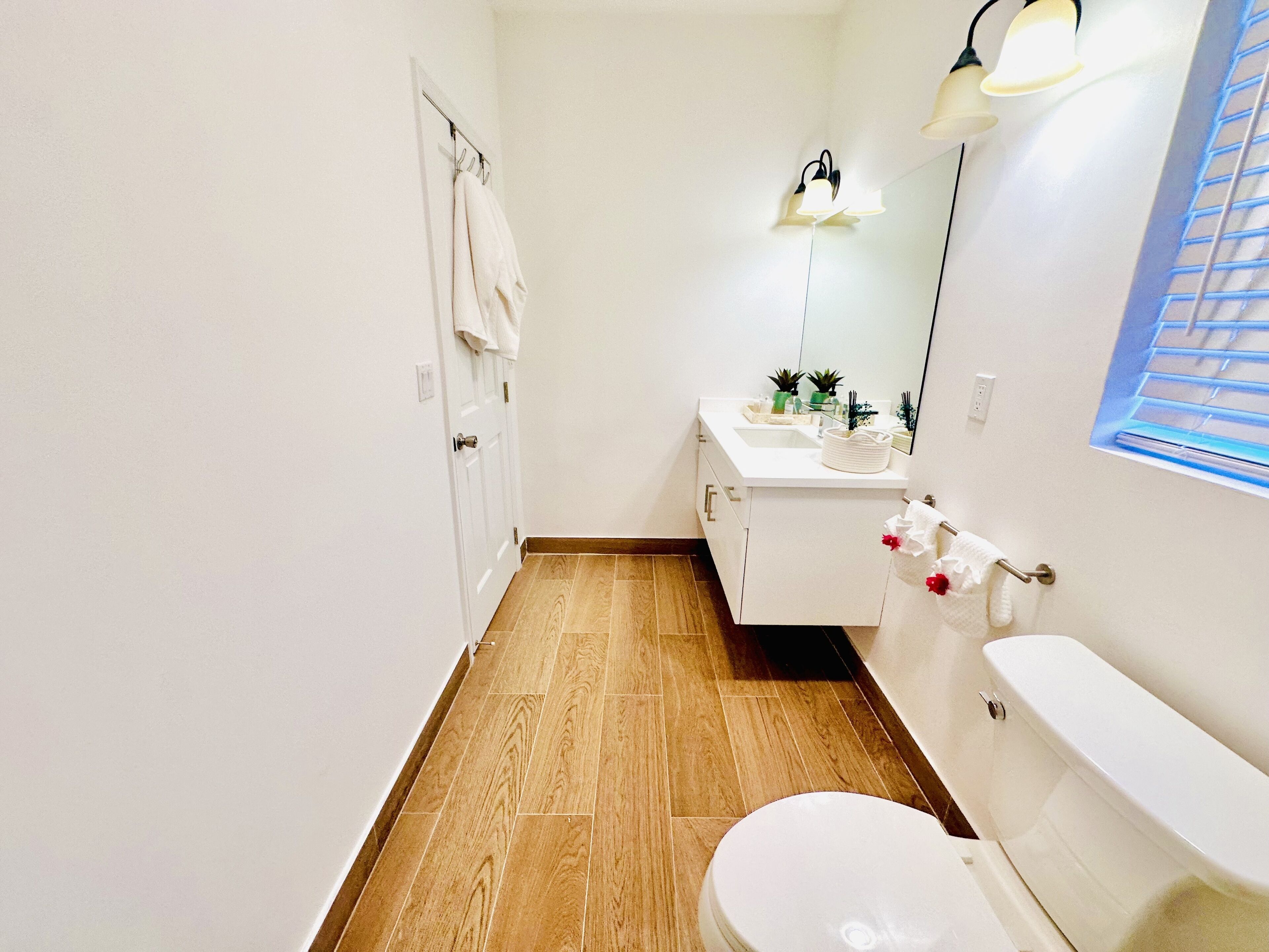 Un baño con inodoro, lavabo y espejo. Hay toallas colgadas en la pared y una ventana con persianas.