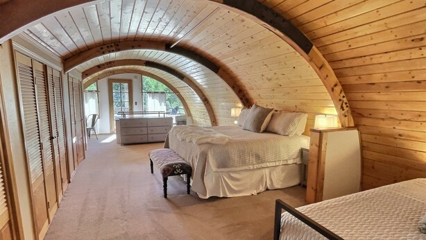 Un dormitorio con techo de madera curvada, una cama con colcha blanca, un banco y una mesita de noche con lámpara.