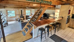 Un interior de cabaña espacioso con escalera de madera, zona de estar con sofá y chimenea, cocina con taburetes de bar y un amplio ventanal con vistas al exterior.