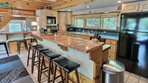 Una cocina moderna con vigas de madera, una isla grande con taburetes de bar, un refrigerador y una ventana con vista a árboles.
