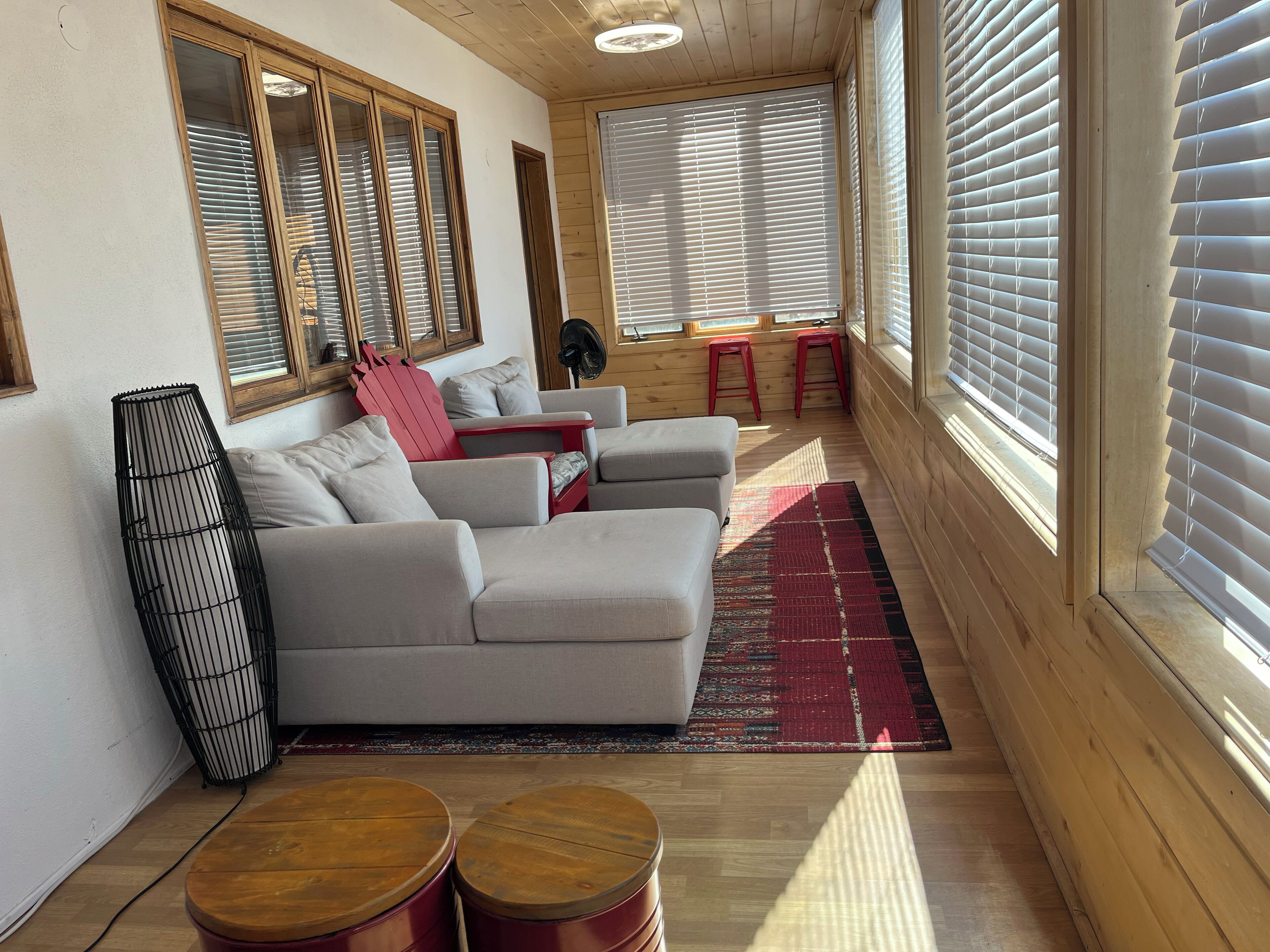 A cozy living room with a sectional sofa, a red chair, two wooden stools, a black floor lamp, and a large window with blinds.