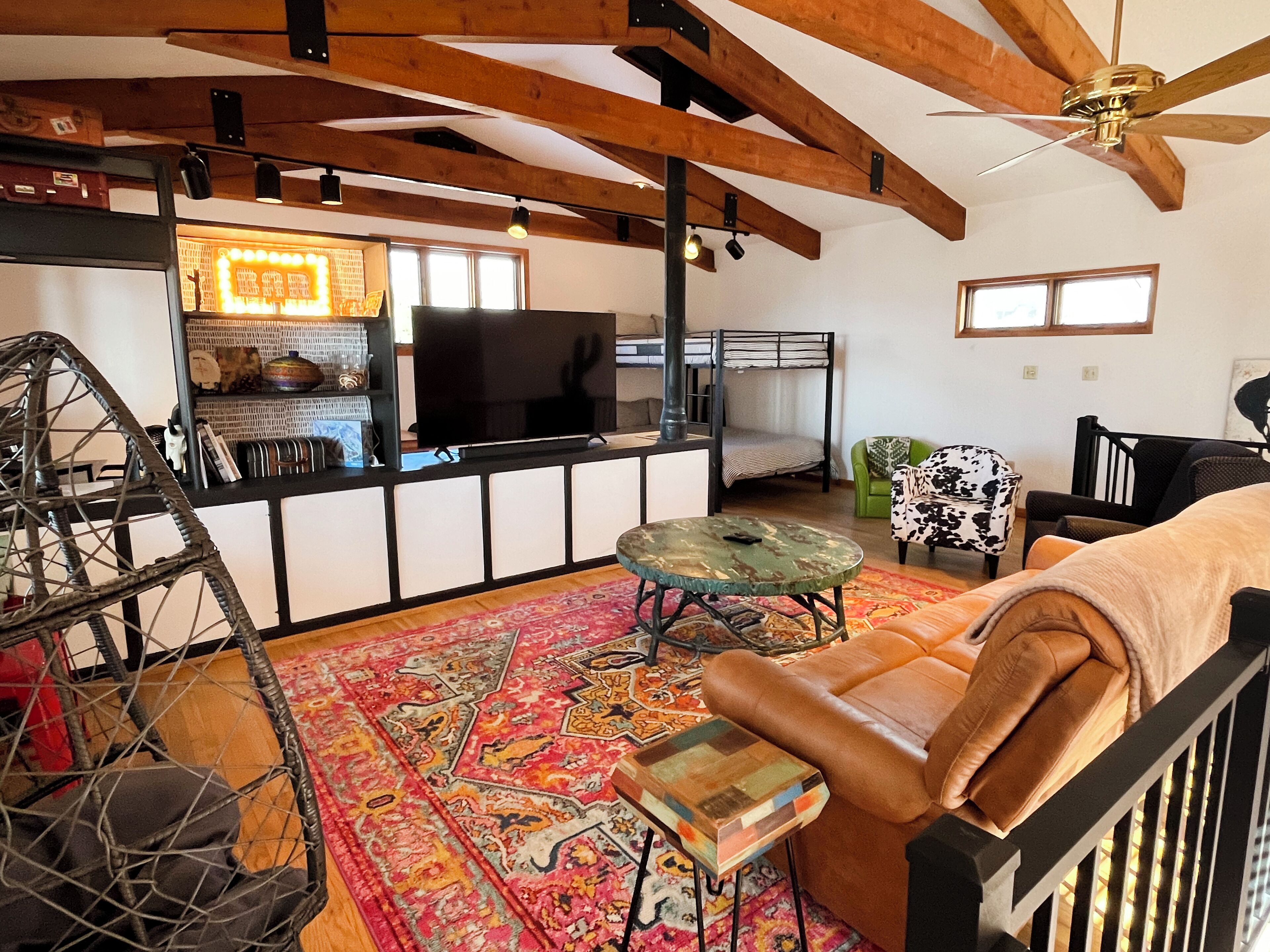 A cozy living room with a high ceiling, exposed wooden beams, a television, a bunk bed, a green armchair, a patterned rug, and a leather sofa.