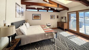 A bedroom with a bed, a nightstand, a dresser, and a window with a view of a snowy landscape. - La Querencia - Taos Foothills Comfort (Arroyo Seco)