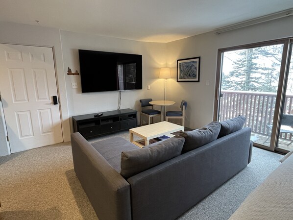 Een moderne woonkamer met een grijze bank, een klein wit tafeltje, een aan de muur bevestigde televisie, een lamp en uitzicht op een terras door een schuifdeur van glas.